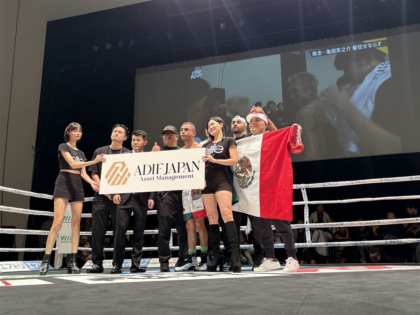 ボクシングのリング上で ADIF JAPAN の幕を掲げるモデルと選手